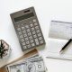 A top-down view image of a calculator on a white table. To the right of the calculator is a notepad with a pen on top of it. To the left of the calculator, there is a small succulent in a pot. Below the calculator is a stack of US one-hundred dollar bills.